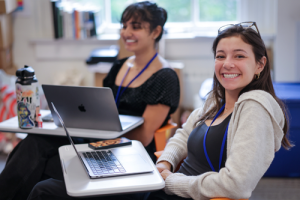 Two Summer students in a class room, open laptops on their desks, smiling.